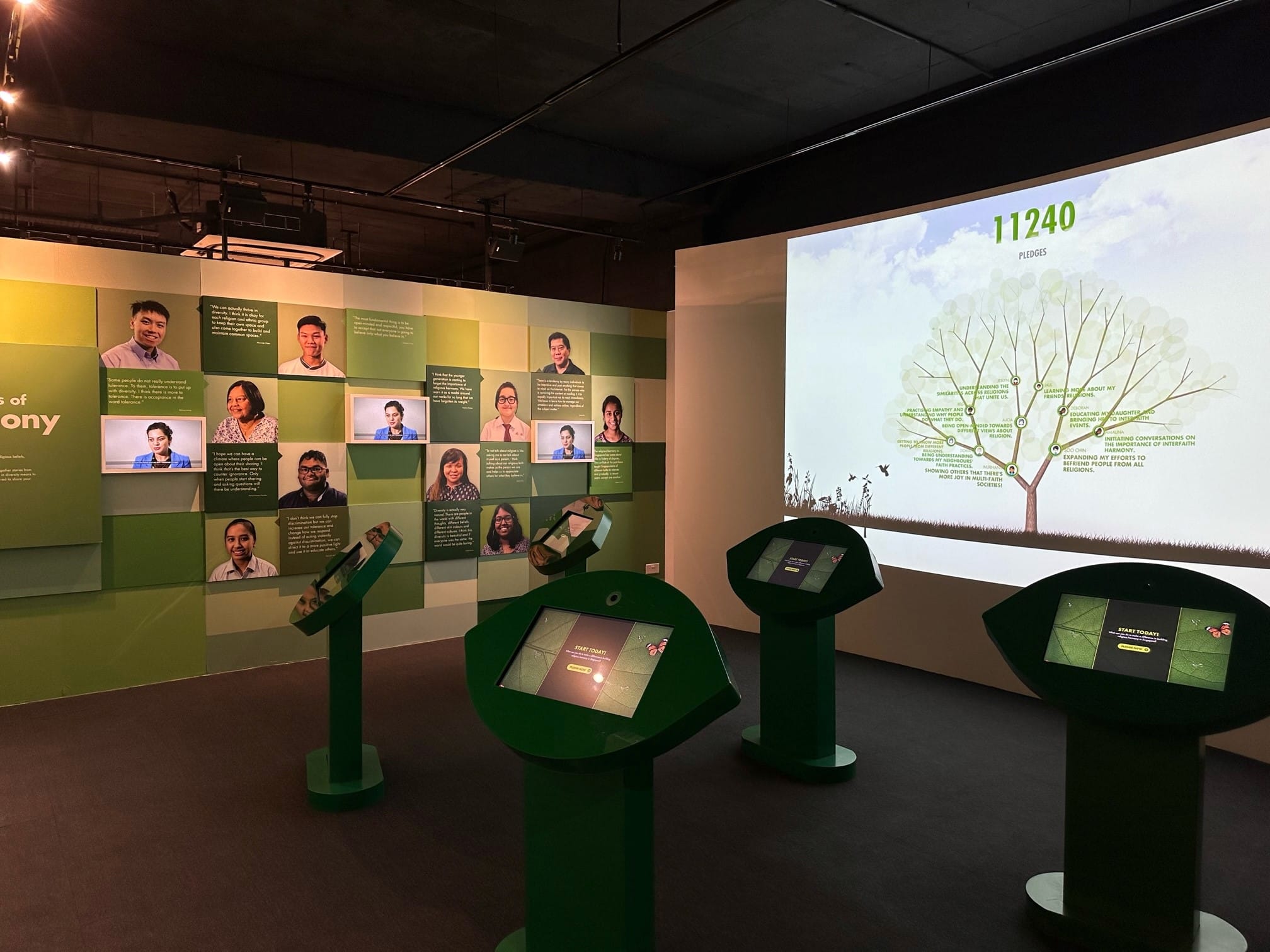 Museum exhibit with green interactive kiosks, a portrait wall, and a projected tree showing 11240 pledges.